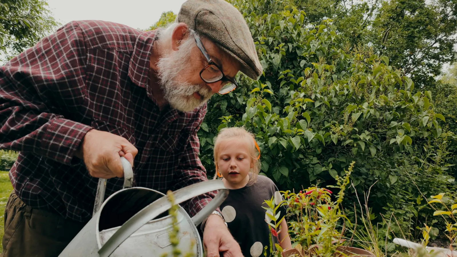 An older gardener showing a young girl how to water plants in a leafy garden.